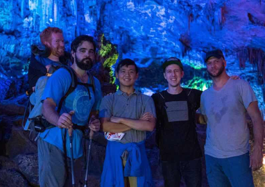 Kevan explore la grotte de la Flûte de roseau à Guilin, en Chine, avec quatre de ses amis. Kevan est porté dans un sac à dos spécial par l’un ses compagnons, qui sont tous les uns à côté des autres et regardent directement la caméra. 