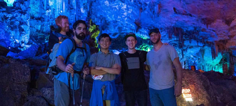 Kevan explores the Reed Flute Caves in Guilin, China with four of his friends. Kevan is being carried in his backpack by one of his companions as they all stand beside each other looking straight into the camera.