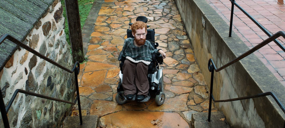 Kevan est assis dans son fauteuil roulant au pied d’un escalier, sans possibilité de se rendre au sommet de celui-ci.