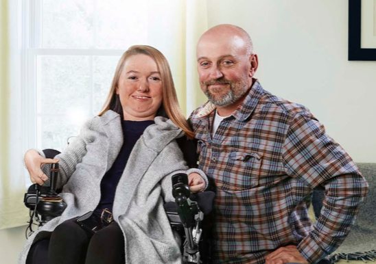 Brianna poses in her wheelchair. Her Dad, who is also her primary caregiver kneels beside her; both are smiling at the camera with sunlight streaming in from a window in the living room.