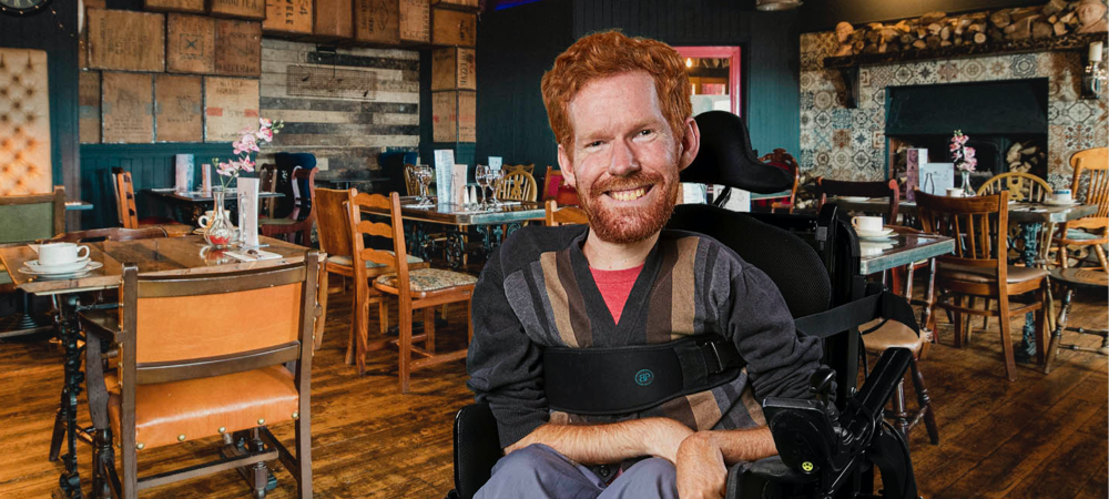 Kevan sits in his wheelchair at his favourite local coffee shop. The image shows him smiling in the middle of the shop with wooden furniture, table flowers and warm lighting.