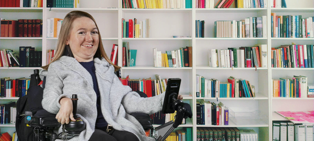Brianna pose dans son fauteuil roulant, devant une bibliothèque remplie de centaines de livres. Elle sourit en regardant directement la caméra.