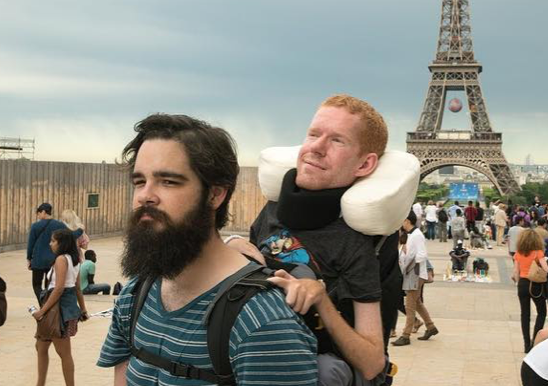 Kevan being carried in his backpack by his friend, Tom, while on a trip in Paris. The Eiffel Tower, a popular tourist landmark can be seen behind them, with tourists milling around.