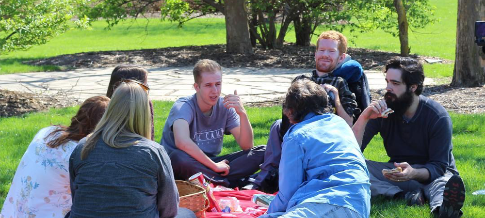 Kevan relaxes in an outdoor picnic, surrounded by greenery and trees. He sits in his wheelchair flanked by two friends.