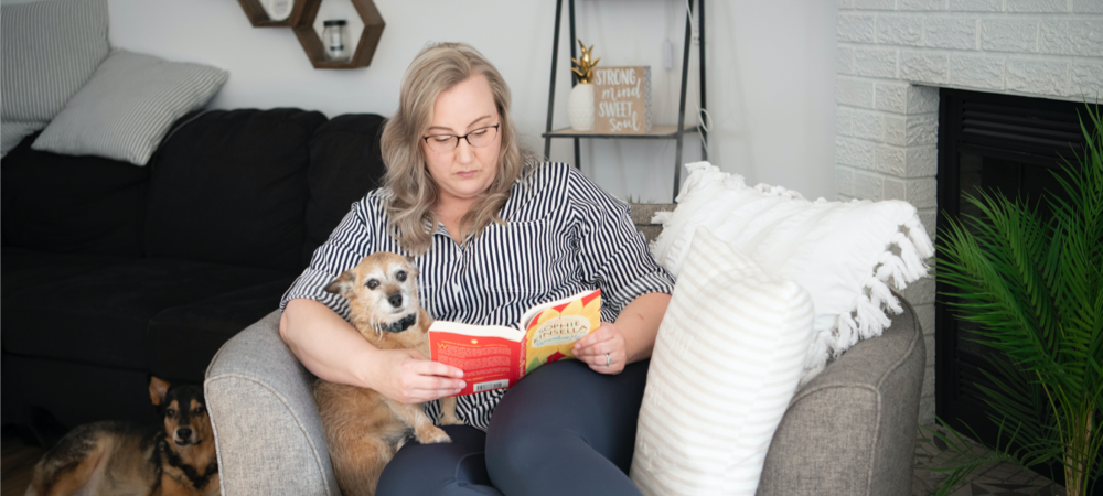 Une mère est assise dans un fauteuil gris avec un chien sur ses genoux, et lit un roman. Un deuxième chien est alongé par terre à côté d’eux.