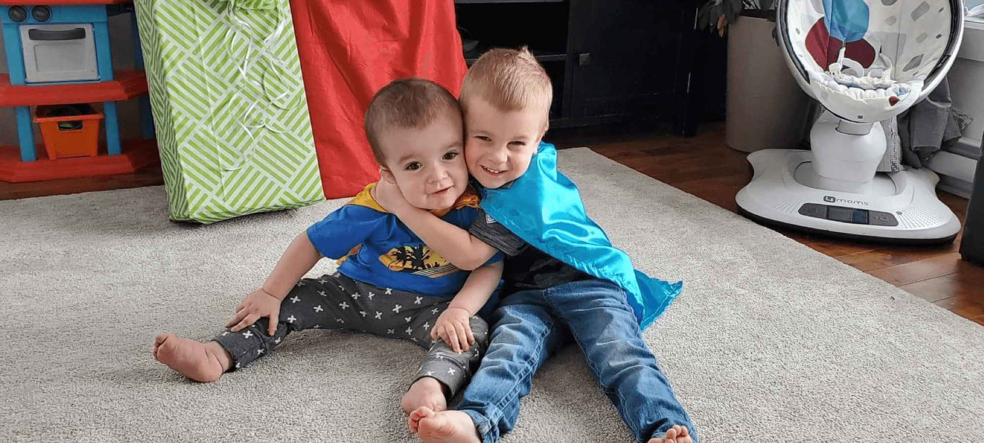 two young boys sitting side-by-side, hugging while wearing superhero capes.