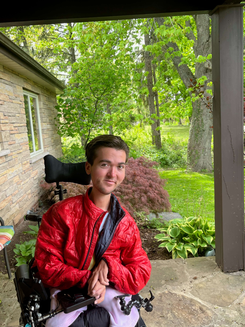 Jeune homme en fauteuil roulant portant un manteau rouge assis sur le porche en pierre d’une maison
