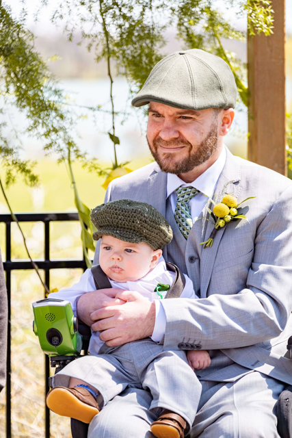Father with SMA wearing gray suit and yellow flower seated in wheelchair holding infant