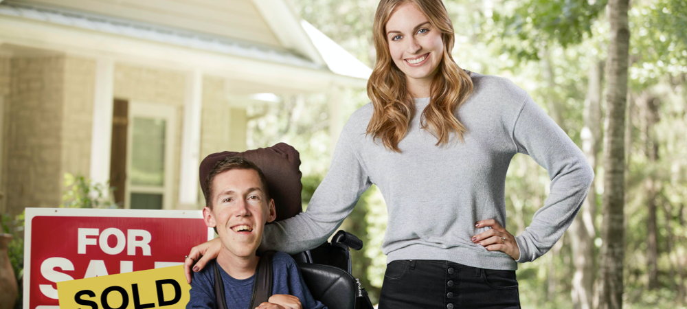 Shane and Hannah smile in front of their newly purchased home with a sold sign next to them. Shane sits in his wheelchair and Hannah stands beside him with her hand on his shoulder.