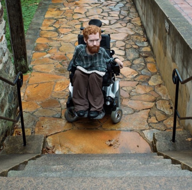 Kevan est assis dans son fauteuil roulant au pied d’un escalier, sans possibilité de se rendre au sommet de celui-ci.