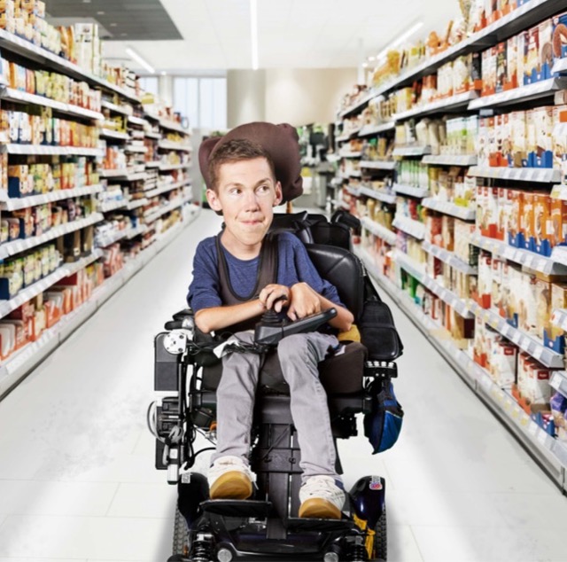 Shane est assis dans son fauteuil roulant, au milieu d’une allée de supermarché remplie de marchandises.