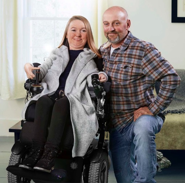 Brianna poses in her wheelchair. Her Dad, who is also her primary caregiver kneels beside her; both are smiling at the camera with sunlight streaming in from a window in the living room.