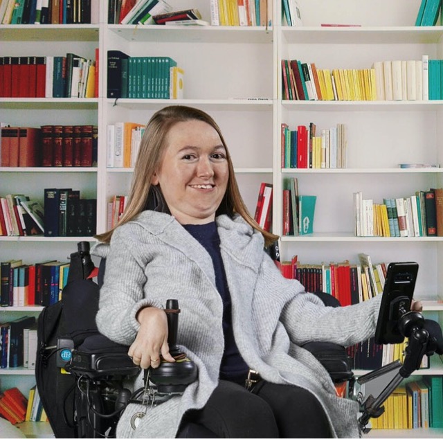 Brianna pose dans son fauteuil roulant, devant une bibliothèque remplie de centaines de livres. Elle sourit en regardant directement la caméra.