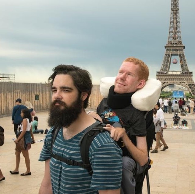 Kevan being carried in his backpack by his friend, Tom, while on a trip in Paris. The Eiffel Tower, a popular tourist landmark can be seen behind them, with tourists milling around.