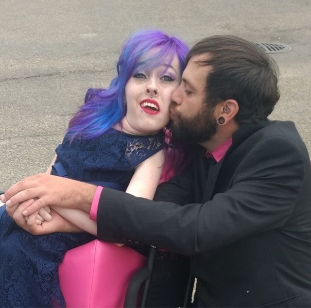 Karli sitting in a wheelchair wearing a blue evening dress. A man kisses her cheek