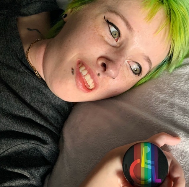Close up of Karli’s head on a pillow. She holds a button pin that shows a rainbow wheelchair icon