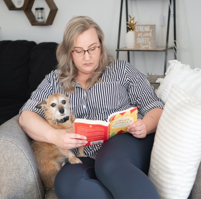 Une mère est assise dans un fauteuil gris avec un chien sur ses genoux, et lit un roman. Un deuxième chien est alongé par terre à côté d’eux.