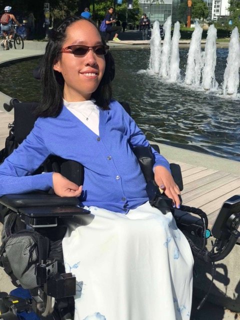 Jeune femme atteinte d’AS portant des lunettes de soleil et un cardigan bleu près d’une fontaine par une journée ensoleillée