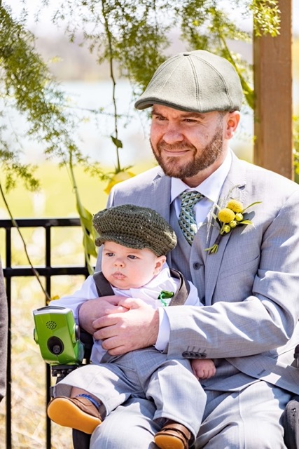 Un père atteint d’AS portant un complet gris et une fleur jaune à sa boutonnière assis dans un fauteuil roulant et tenant un bébé